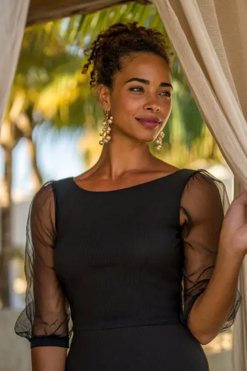 A confident woman in a fitted black midi dress with sheer sleeves and pearl earrings, standing beneath linen curtains swaying at a tropical rehearsal dinner setup