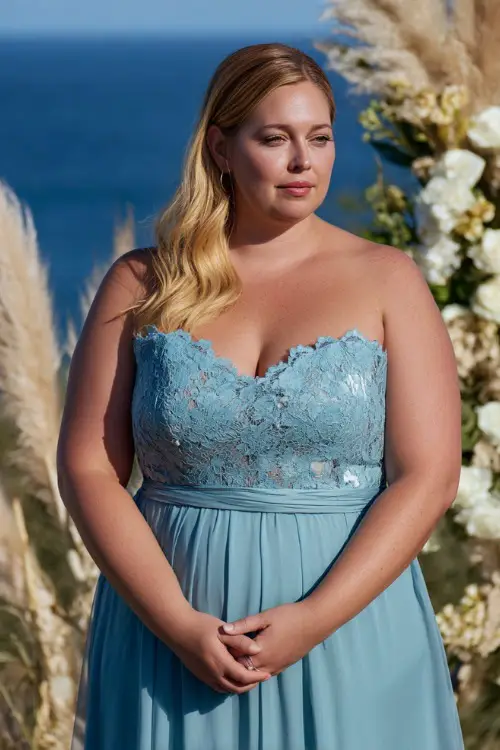 A plus-size woman in a soft blue midi dress with a sweetheart neckline and lace overlay, standing at a coastal wedding altar surrounded by sea grasses and white blooms 