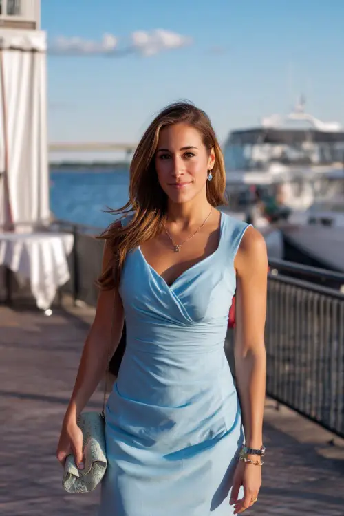 A stylish woman in a sleeveless baby blue dress with a softly draped neckline and clutch, walking near a waterfront rehearsal dinner location 