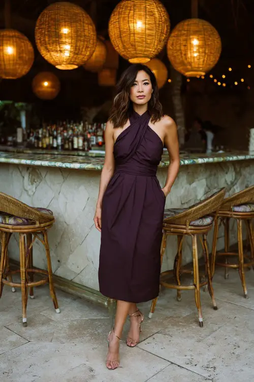 A woman in a dark plum sleeveless midi dress with elegant draping and strappy heels, standing near a rehearsal dinner bar surrounded by rattan lanterns and soft lighting 