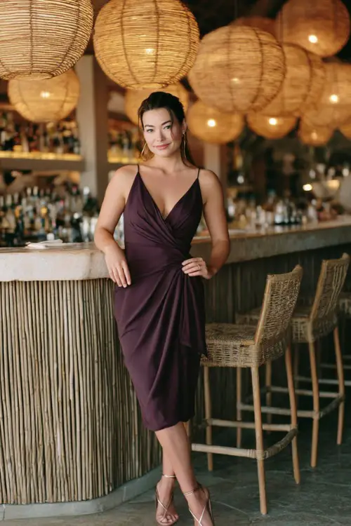 A woman in a dark plum sleeveless midi dress with elegant draping and strappy heels, standing near a rehearsal dinner bar surrounded by rattan lanterns and soft lighting 