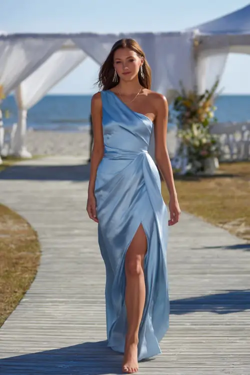 A woman in a light blue one-shoulder satin gown with a high slit and elegant earrings, walking across a wooden boardwalk toward a beachside ceremony setup