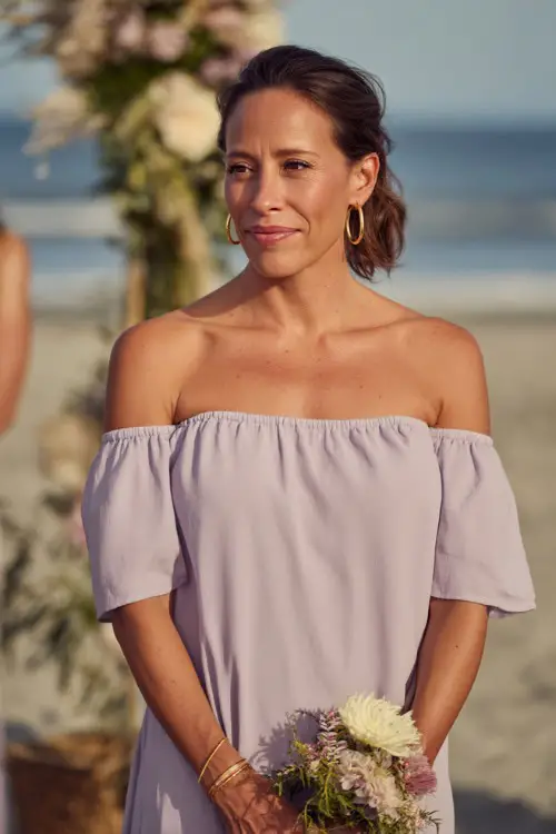 A woman in a light lavender off-the-shoulder dress with a relaxed fit and gold hoop earrings, standing beside low floral arrangements at a casual beach ceremony