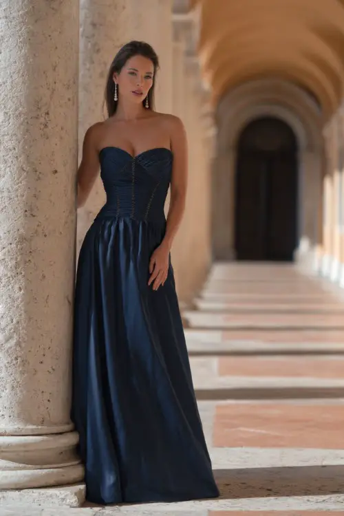 A woman in a navy blue floor-length satin gown with a structured bodice and pearl drop earrings, standing beneath an archway at a grand summer wedding venue