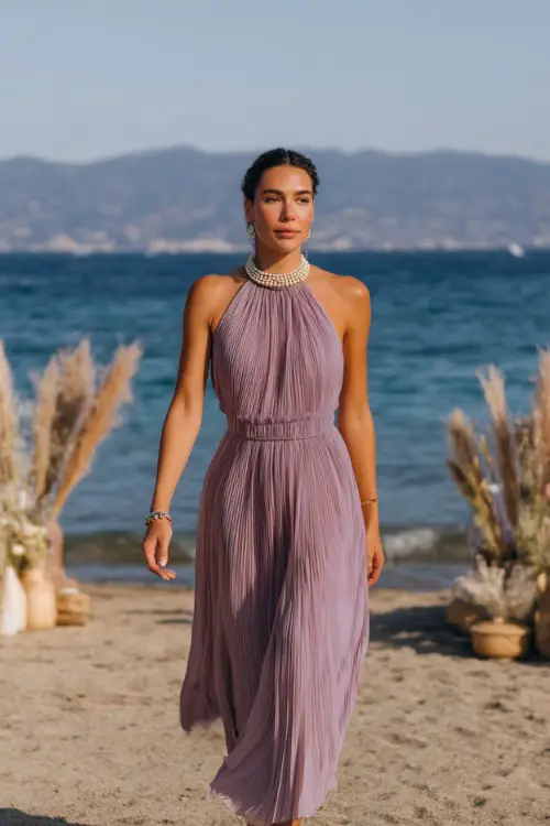 A woman in a pastel lavender pleated midi dress with a high neckline and pearl accessories, walking along the shoreline toward a boho-style beach wedding setup