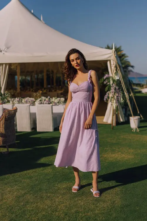 A woman in a pastel lavender smocked midi dress with shoulder ties and flat sandals, standing beside a white tented reception setup on the grass
