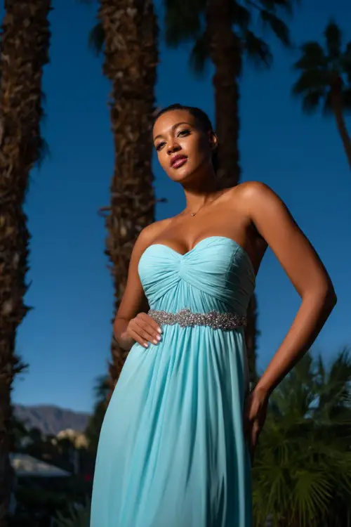 A woman in a powder blue floor-length gown with a sweetheart neckline and jeweled waistband, posing beneath tall palm trees at a sunset destination black-tie event 