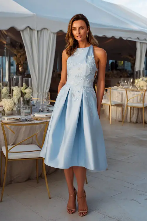 A woman in a sky blue fit-and-flare midi dress with embroidered details and heels, standing near a white tented reception space with elegant table settings 