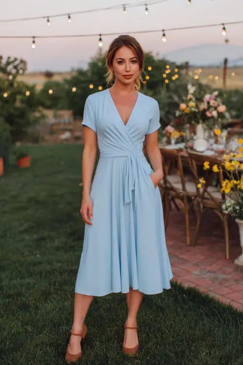 A woman in a sky blue wrap dress with short sleeves and tan heels, standing near a backyard wedding setup with fairy lights and florals 