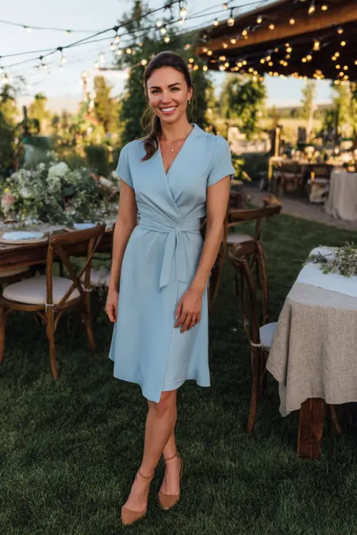 A woman in a sky blue wrap dress with short sleeves and tan heels, standing near a backyard wedding setup with fairy lights and florals 