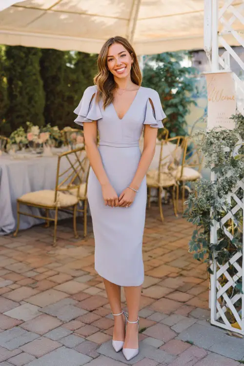 A woman in a soft lavender tea-length dress with short flutter sleeves and strappy heels, standing near a welcome sign at an elegant backyard rehearsal dinner