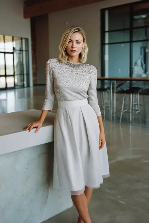 A woman in a soft silver midi dress with a beaded neckline and light chiffon skirt, standing near a modern rehearsal dinner space with clean minimalist décor 