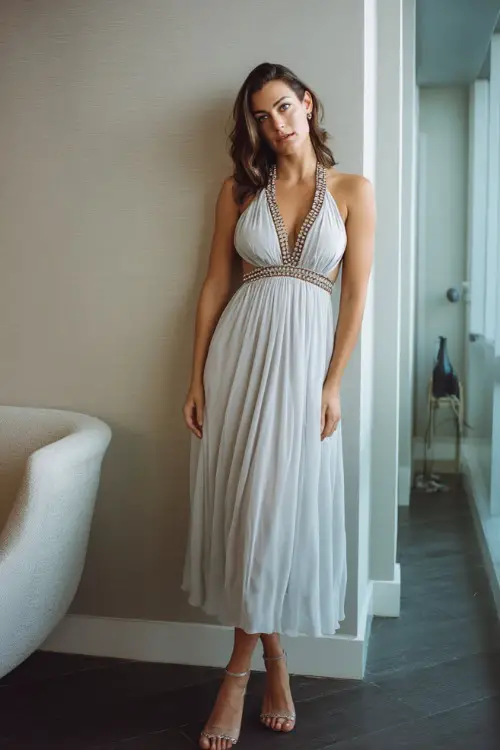 A woman in a soft silver midi dress with a beaded neckline and light chiffon skirt, standing near a modern rehearsal dinner space with clean minimalist décor 