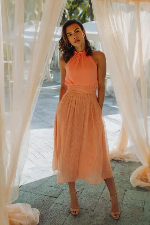 A woman wearing a coral sleeveless dress with a pleated midi skirt and neutral heels, standing under sheer fabric draping at a destination rehearsal dinner