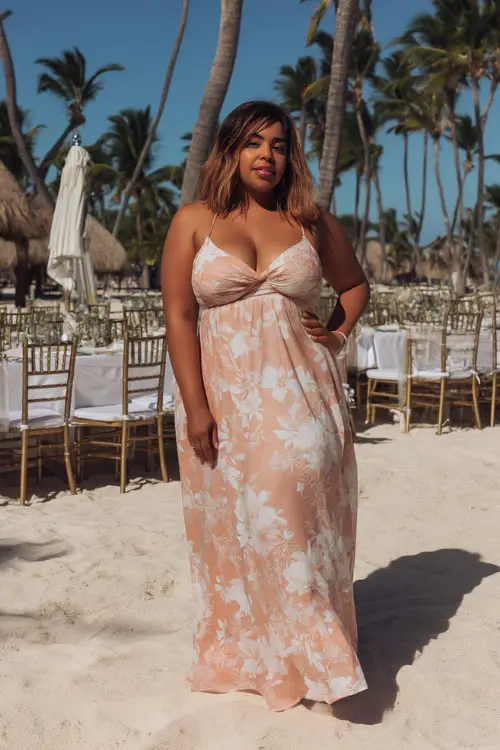 A woman with a curvy figure in a peach midi dress with tie straps and white floral prints, standing at a beach wedding setup with palm trees and linen-draped chairs 