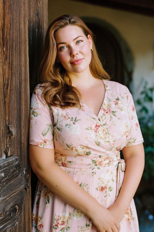 plus size woman standing in a rustic garden entrance, wearing a light pink floral fit and flare dress with short sleeves and a cinched waist, soft romantic hairstyle