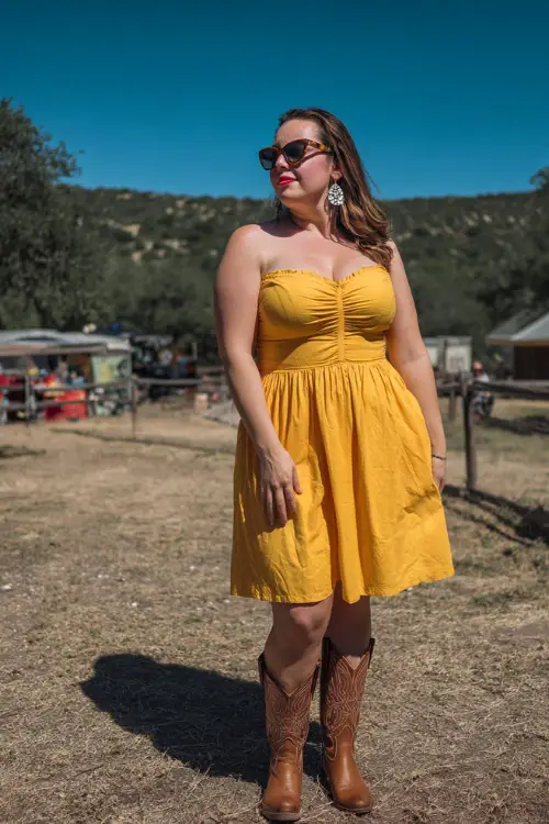 A plus-size woman wearing a smocked midi sundress in sunflower yellow with classic western boots, styled with statement earrings and sunglasses 
