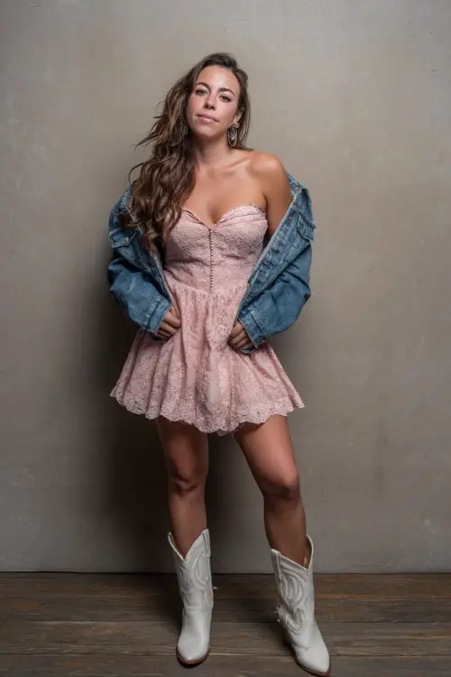 A woman in a blush pink lace dress with flutter sleeves, paired with white cowboy boots and a denim jacket draped over her shoulders