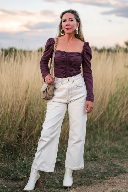 A woman over 40 in wide-leg jeans, a ruched square-neck blouse, and white cowboy boots, accessorized with a small leather purse and hoop earrings 