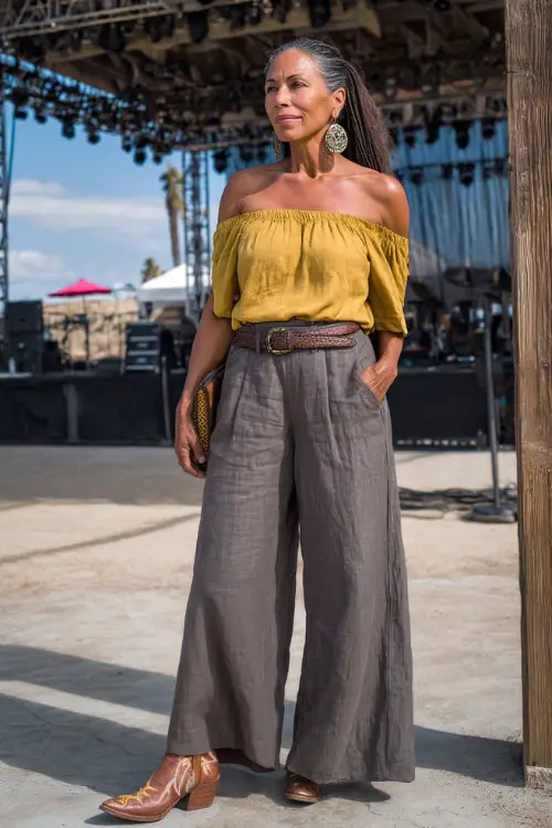 A woman over 40 styled in wide-leg linen pants, a fitted off-shoulder blouse, and tall embroidered cowboy boots, accessorized with boho earrings and a leather clutch 