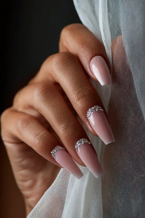Bridal hand posing against a sheer curtain, extra-long coffin-shaped acrylic nails in pale pink with crystal bead clusters along the tips 