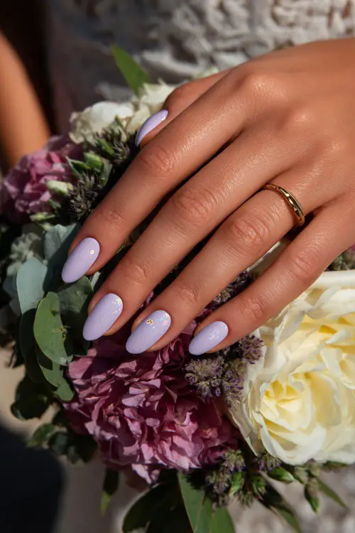 Bride’s fingers curled around a floral bouquet, almond nails in pastel lilac with shimmer overlay and tiny white floral stamp on the ring finger 