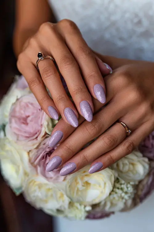Bride’s fingers curled around a floral bouquet, almond nails in pastel lilac with shimmer overlay and tiny white floral stamp on the ring finger