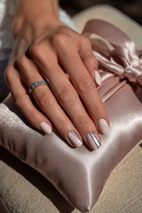 Bride’s hand beside a silk ring pillow, square nails in soft rose-beige polish with silver chrome striping on two accent nails 