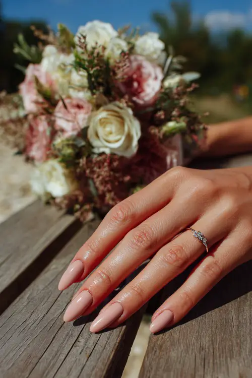 Bride’s hand beside bouquet on a picnic table, almond nails in soft rose beige with floral stamping on each tip, outdoor summer wedding light 