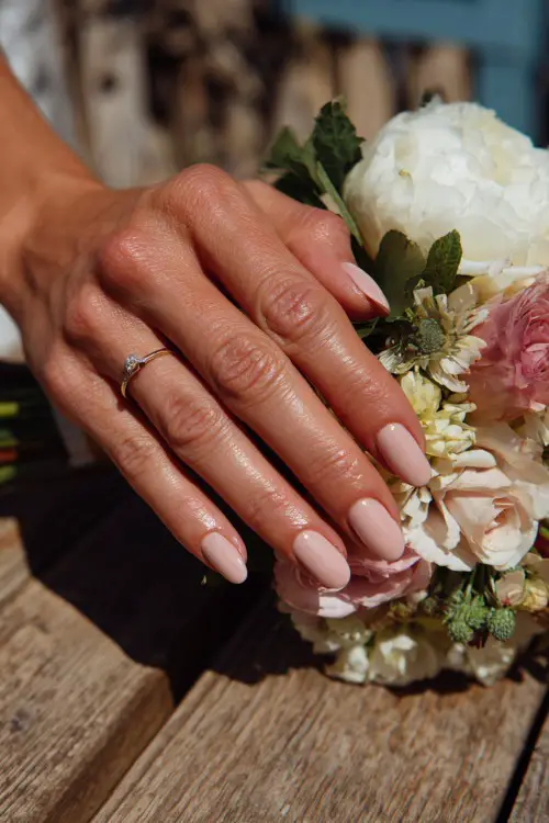 Bride’s hand beside bouquet on a picnic table, almond nails in soft rose beige with floral stamping on each tip, outdoor summer wedding light