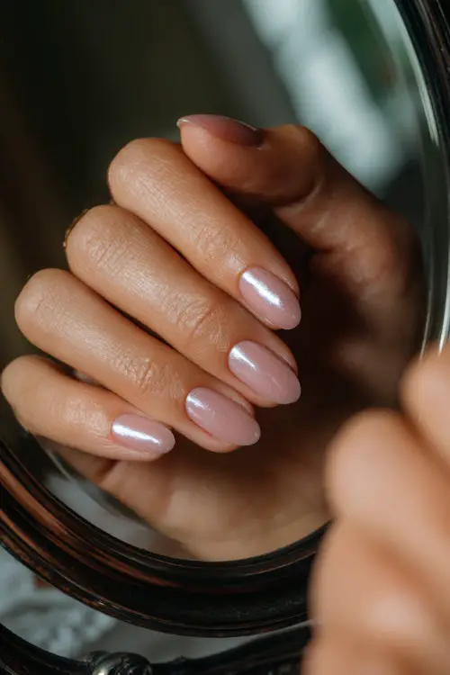 Bride’s hand brushing over mirror, short oval nails in glassy pastel pink with chrome shimmer tip on one nail, classic vanity background 