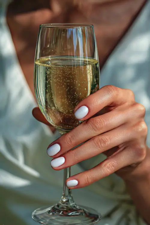 Bride’s hand holding a champagne glass, nails painted in white ombré with subtle sparkle, elegant party background blur 