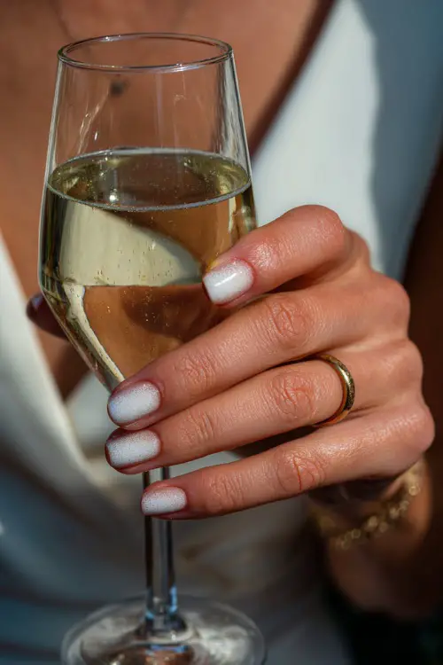 Bride’s hand holding a champagne glass, nails painted in white ombré with subtle sparkle, elegant party background blur 