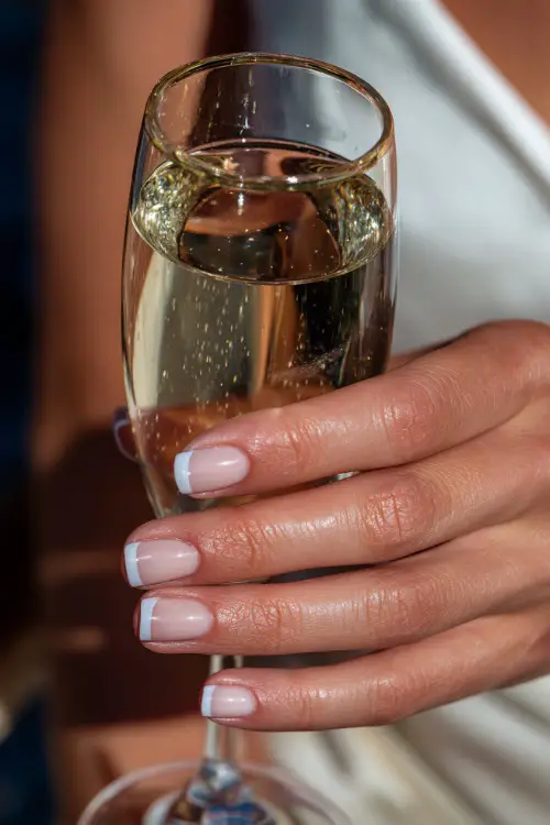Bride’s hand holding a crystal champagne flute, nails in classic French manicure with ultra-thin white tips and a glossy finish, elegant reception background blur 