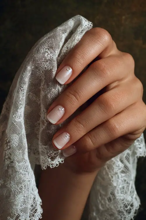 Bride’s hand holding a lace handkerchief, short square nails in sheer white with subtle shimmer and micro pearl studs, romantic vintage background
