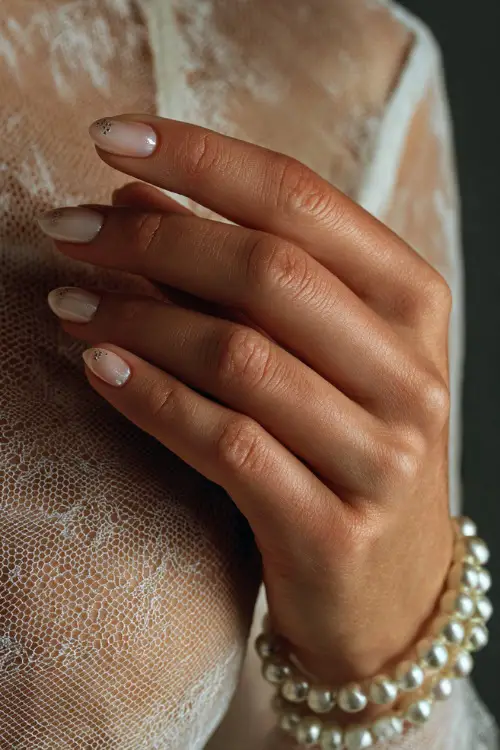 Bride’s hand holding a pearl bracelet, almond nails in sheer white with tiny crystal dots at the cuticles, elegant and timeless design