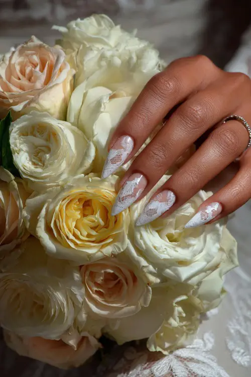 Bride’s hand over bouquet of soft roses, long stiletto acrylic nails with a translucent base and white lace detailing