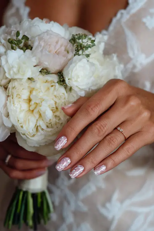 Bride’s hand over soft bouquet, almond nails painted pale pink with white floral stamping and clear crystal details 