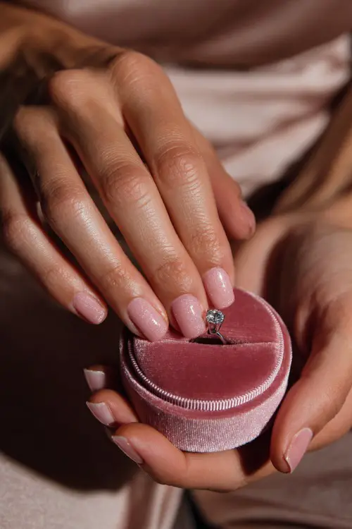 Bride’s hand placing a ring in a velvet box, showcasing light pink sheer jelly nails with fine silver glitter gradient on tips, soft romantic background 