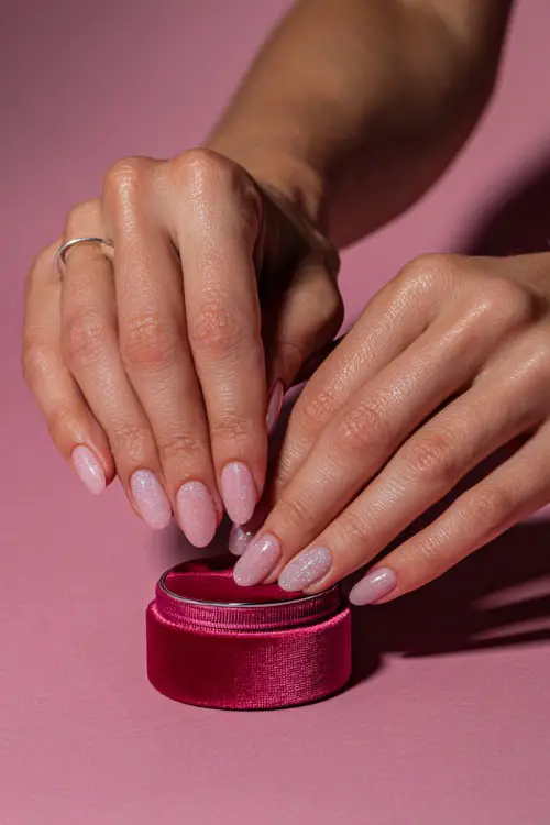 Bride’s hand placing a ring in a velvet box, showcasing light pink sheer jelly nails with fine silver glitter gradient on tips, soft romantic background