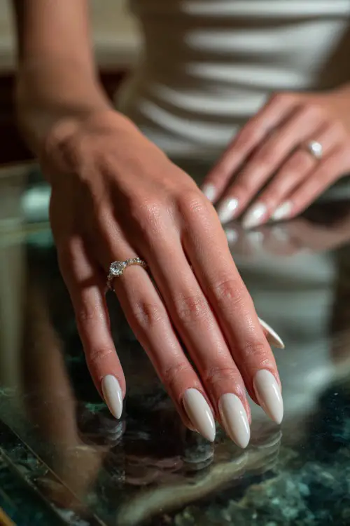 Bride’s hand resting gently on a glass table, showcasing stiletto nails in matte ivory with minimal pearl drop charm on each ring finger 