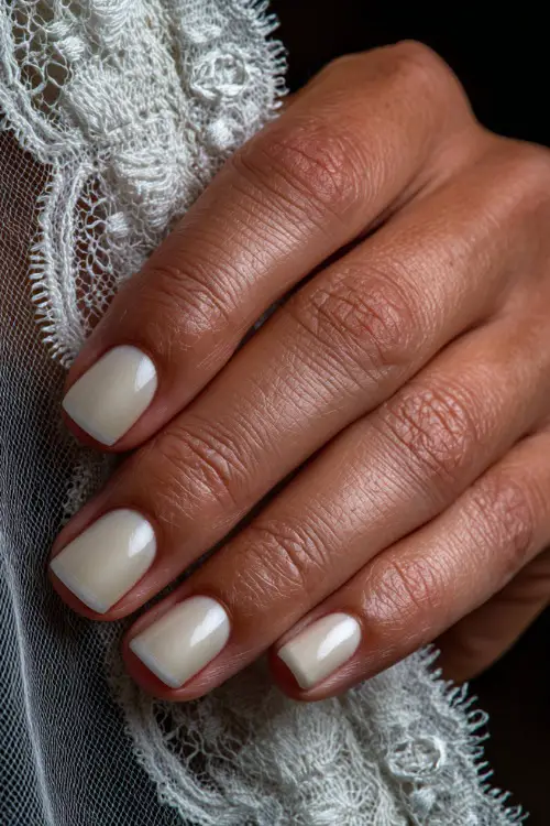 Bride’s hand resting on a lace-trimmed veil, square-shaped nails in creamy white with a chrome French tip and smooth shine 