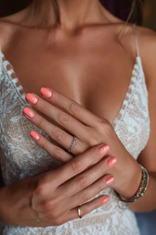 Bride’s hand softly grazing her neckline, square nails painted in pastel coral with chrome glitter on one accent nail 