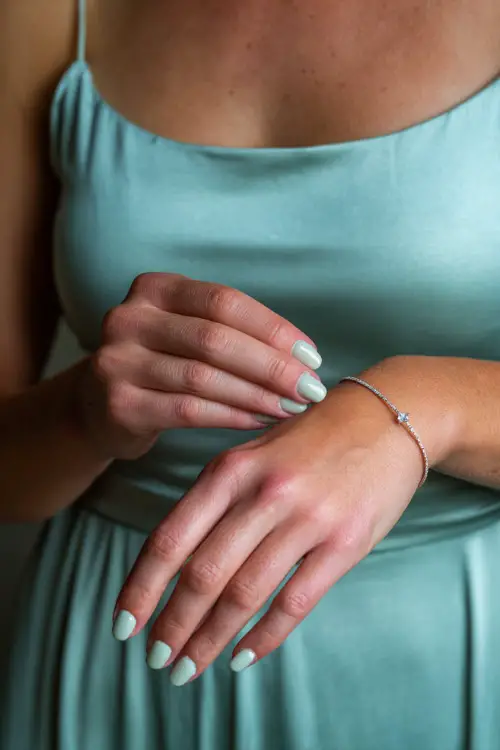 Bridesmaid adjusting her bracelet, rounded nails in pastel sage green with clear gloss, one finger featuring a tiny crystal accent 