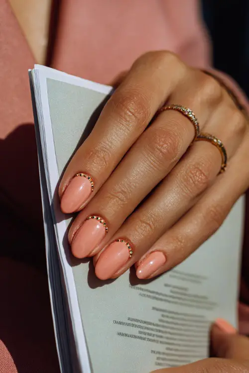 Bridesmaid’s hand gently holding a wedding program, almond nails in pastel peach with glossy finish and small gold beads forming a delicate curve at the base 