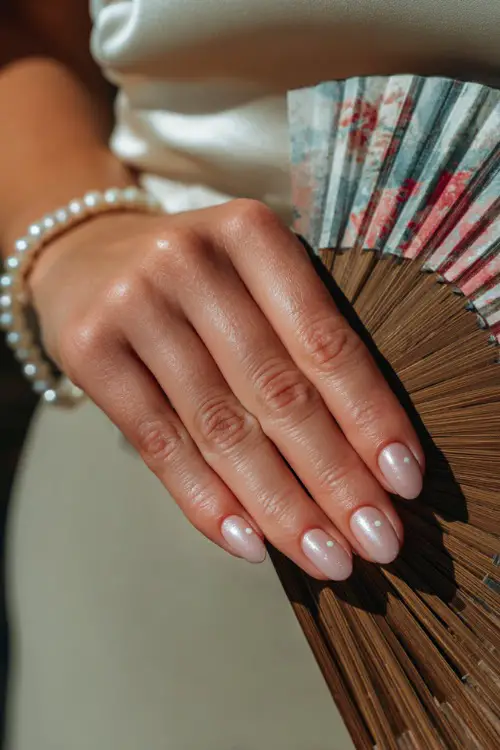 Bridesmaid’s hand resting beside a wedding program fan, oval nails in sheer pastel rose with pearl shimmer and minimalist white dots at the cuticle