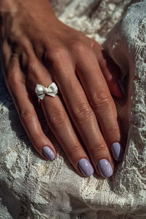 Close-up of a bride’s hand over a vintage lace tablecloth, nails in matte pale lavender with a glossy swirl overlay and small white bow charm on the ring finger 