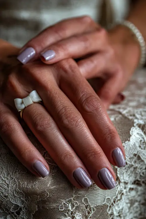 Close-up of a bride’s hand over a vintage lace tablecloth, nails in matte pale lavender with a glossy swirl overlay and small white bow charm on the ring finger 