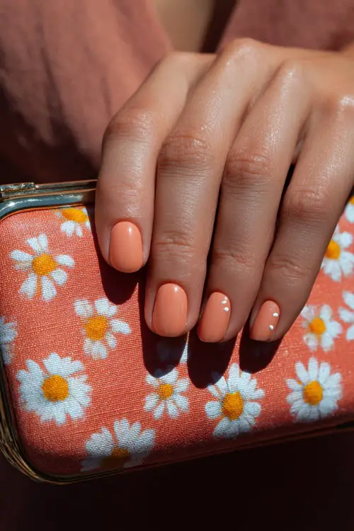 Close-up of a guest’s hand holding a summer floral clutch, short rounded nails in pastel peach with glossy finish and tiny white daisy accents