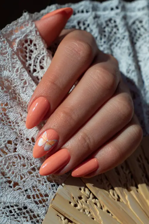 Close-up of a hand resting on a lace fan, almond-shaped nails in pastel coral with a single butterfly decal on the middle finger 